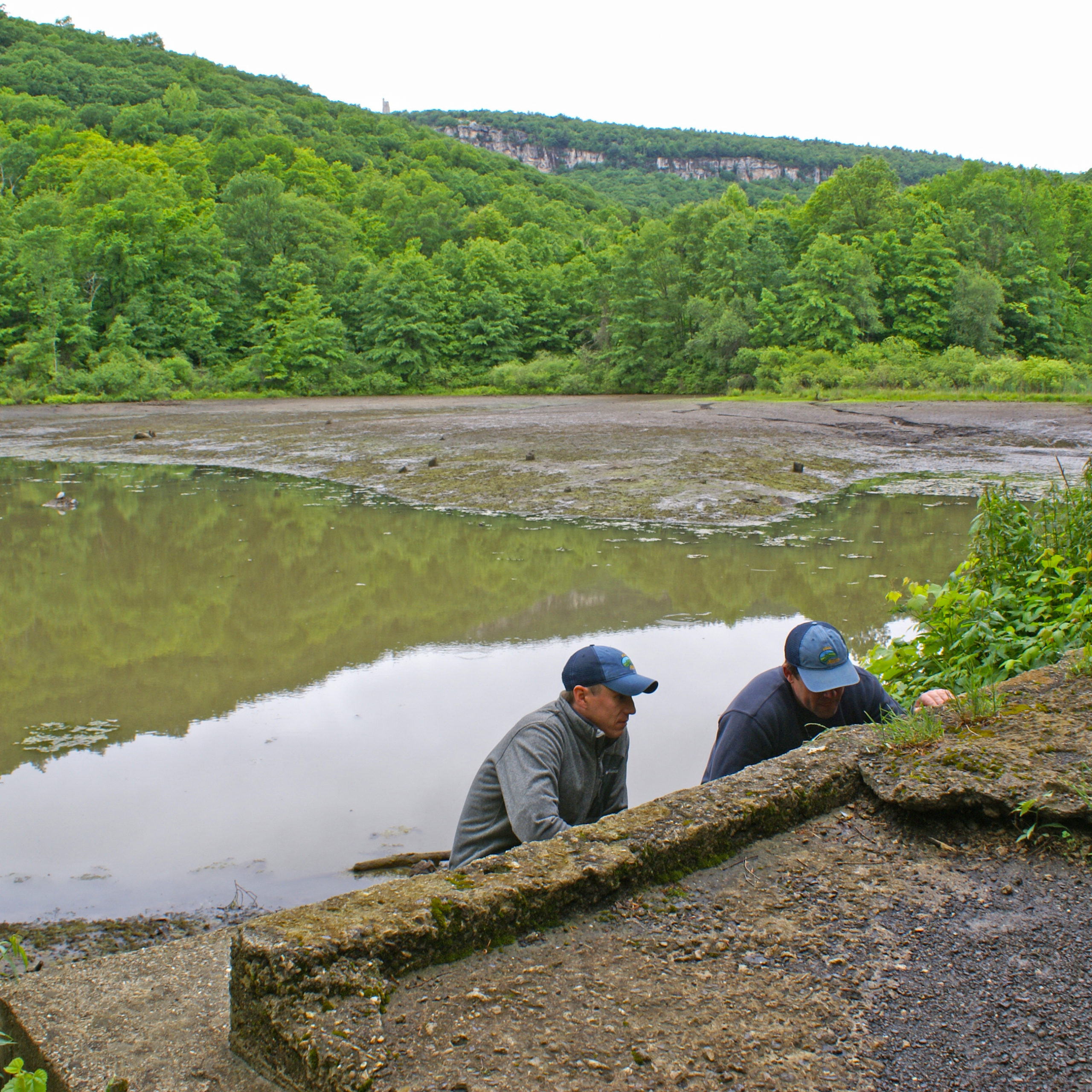 Chuck Reid and Jon Winans investigate Duck Pond Dam in the foreground, behind them Duck Pond drained of most water, Skytop tower and cliff in the background