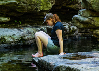 A young person sits down on a large flat rock next to the stream, to feel the temperature of the water at Split Rock.