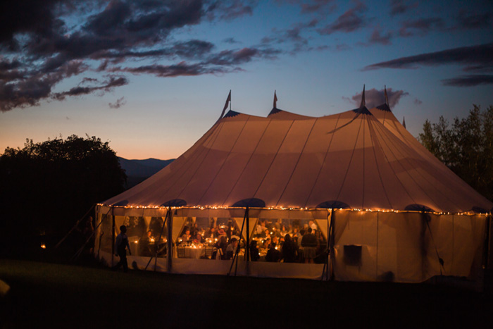 A wedding tent with string lights sits in a meadow overlooking a beautiful sunset