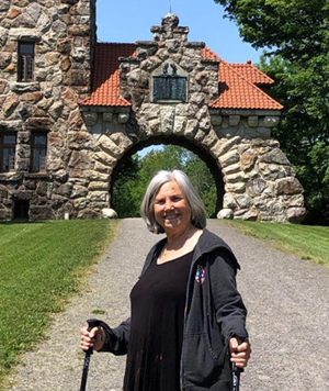 A hiker stands in front of the Mohonk Preserve Testimonial Gateway Tower with hiking poles on a sunny day